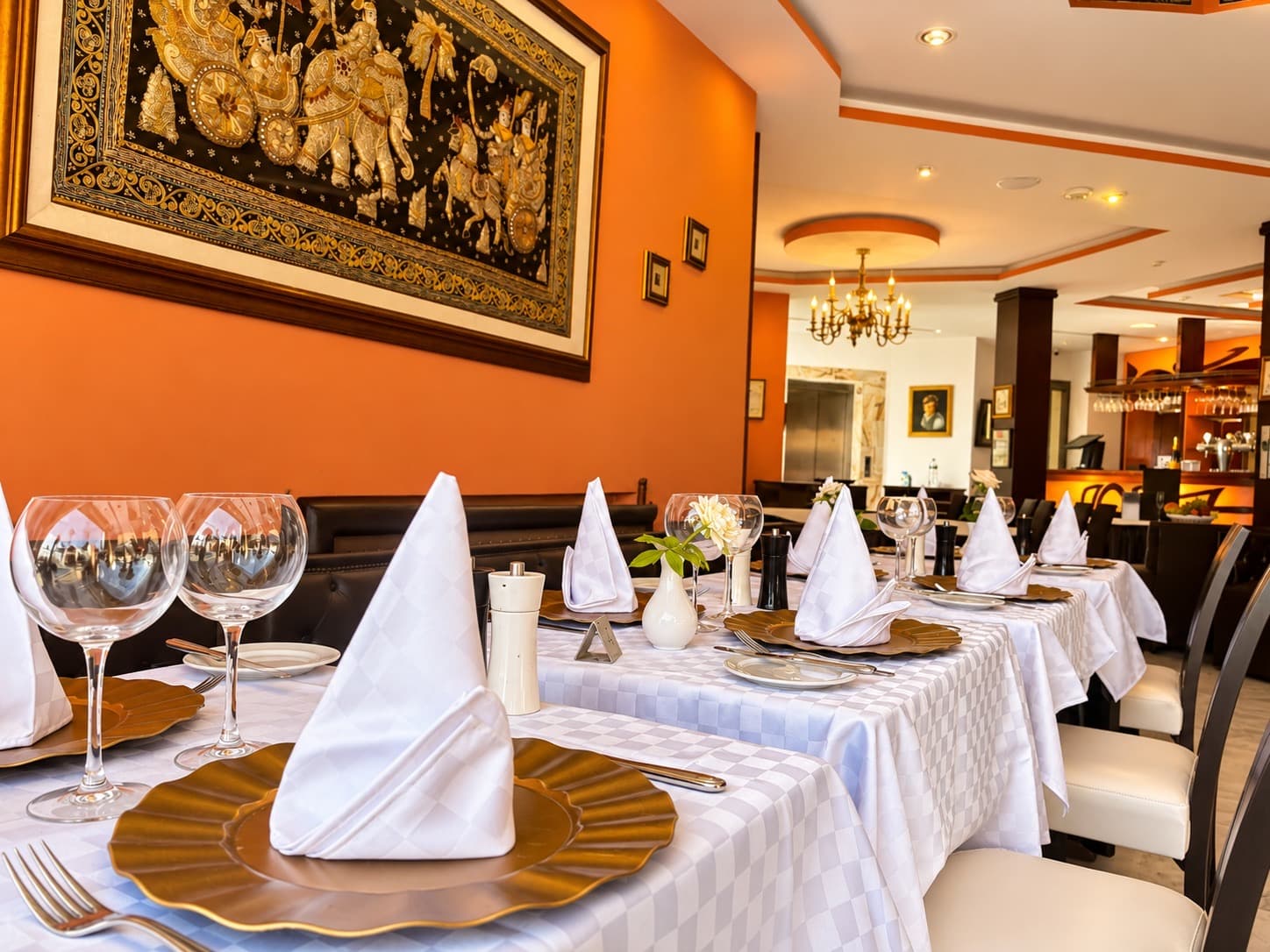 Salle du restaurant aux murs orangés, tables dressées de nappes blanches et assiettes dorées, lustre en laiton